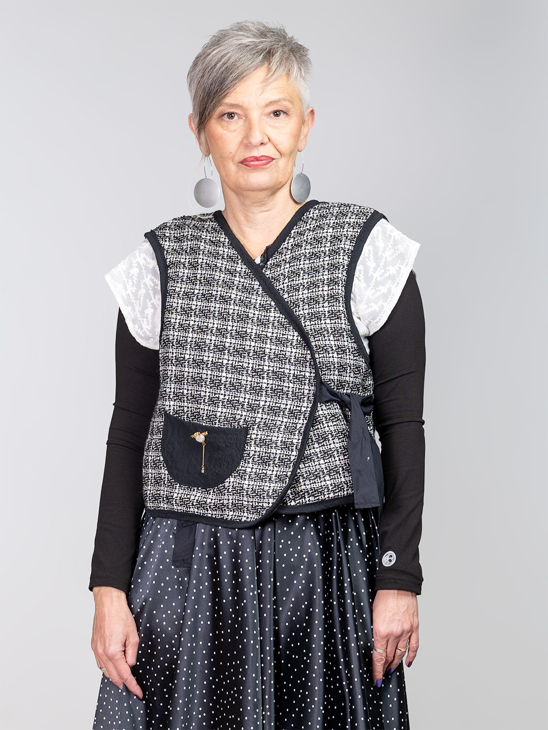 Ženski dvostrani prsluk s crno-bijelim bukle uzorkom, asimetričnim vezanjem i ukrasnim džepom u kombinaciji s tamnom suknjom na točkice.