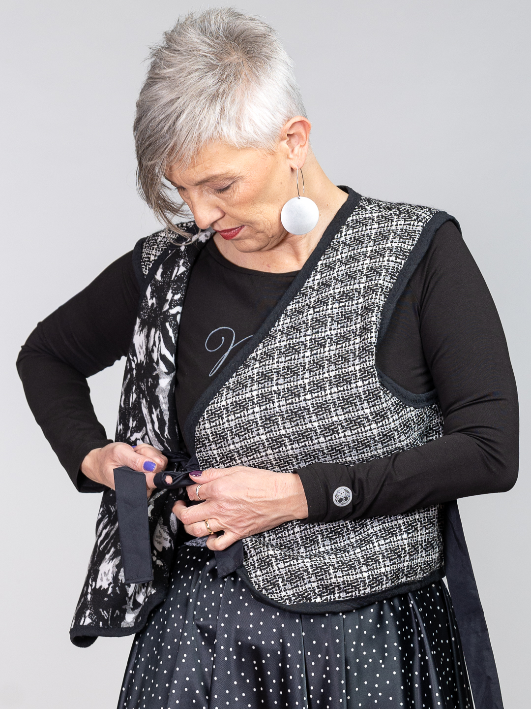 Ženski dvostrani prsluk s crno-bijelim bukle uzorkom, asimetričnim vezanjem i ukrasnim džepom u kombinaciji s tamnom suknjom na točkice.