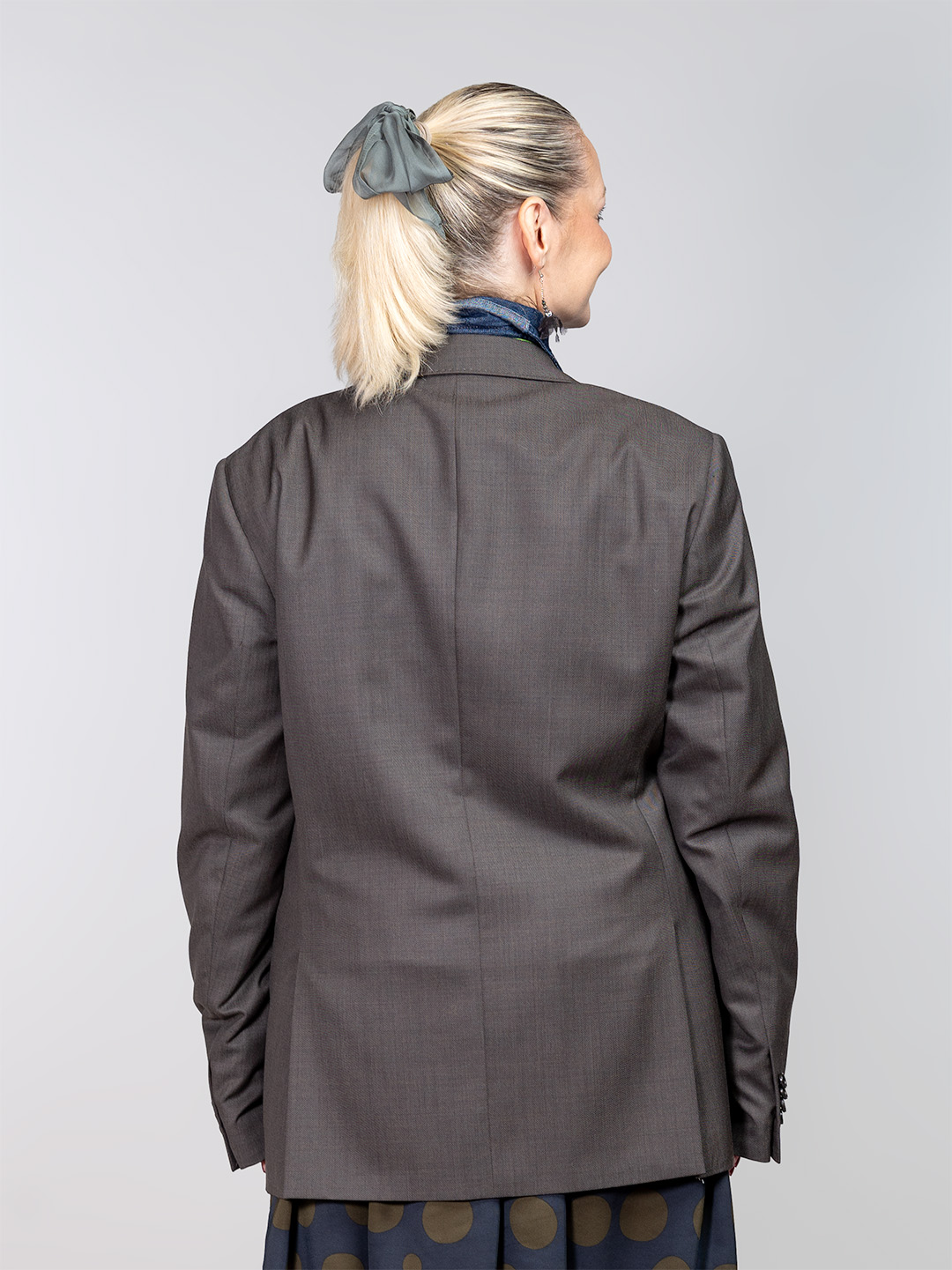 Maslinasti redizajnirani sako Suffragette(6) Žena s osmijehom nosi maslinasti upcycled sako ukrašen tamnom čipkom na reveru i zelenim gumbima, u kombinaciji sa zelenom kravatom i točkastom suknjom.