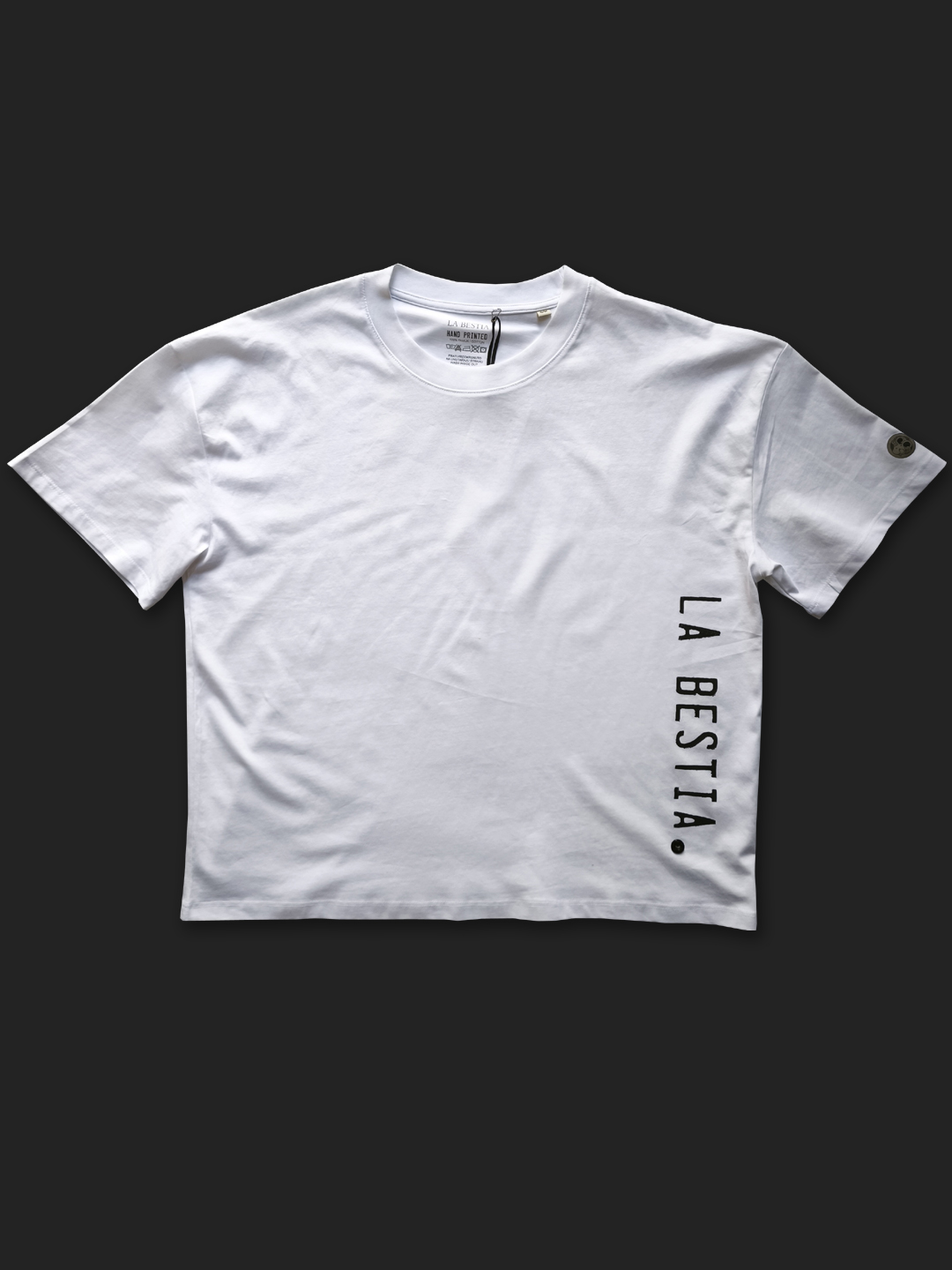 Bijela oversized majica s vertikalnim srebrno-crnim natpisom "LA BESTIA" na bočnoj strani.